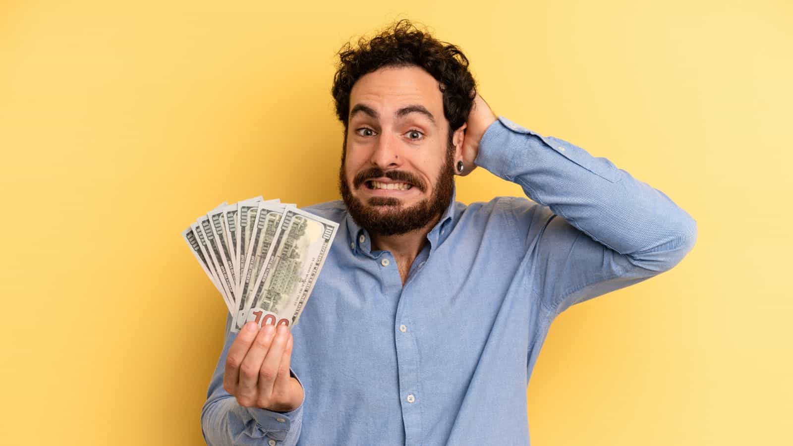 A man with curly hair and a beard, wearing a light blue shirt, holds a fan of U.S. dollar bills in one hand and touches the back of his head with the other, standing against a yellow background.