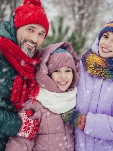 A man, a woman, and a child stand outdoors in winter clothing, smiling at the camera. Snow is falling, and trees covered in snow are visible in the background.
