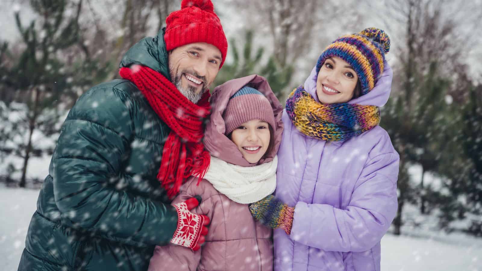 A man, a woman, and a child stand outdoors in winter clothing, smiling at the camera. Snow is falling, and trees covered in snow are visible in the background.