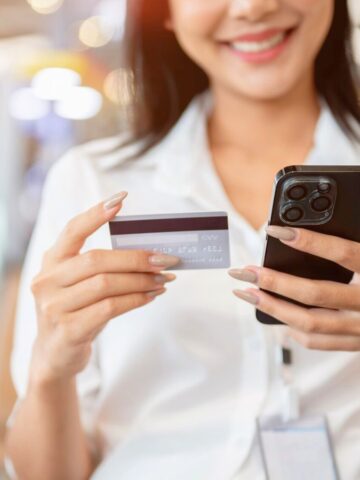 A woman in a white shirt holds a credit card in one hand and a smartphone in the other, smiling while looking at her phone. She is seated indoors with a blurred background.
