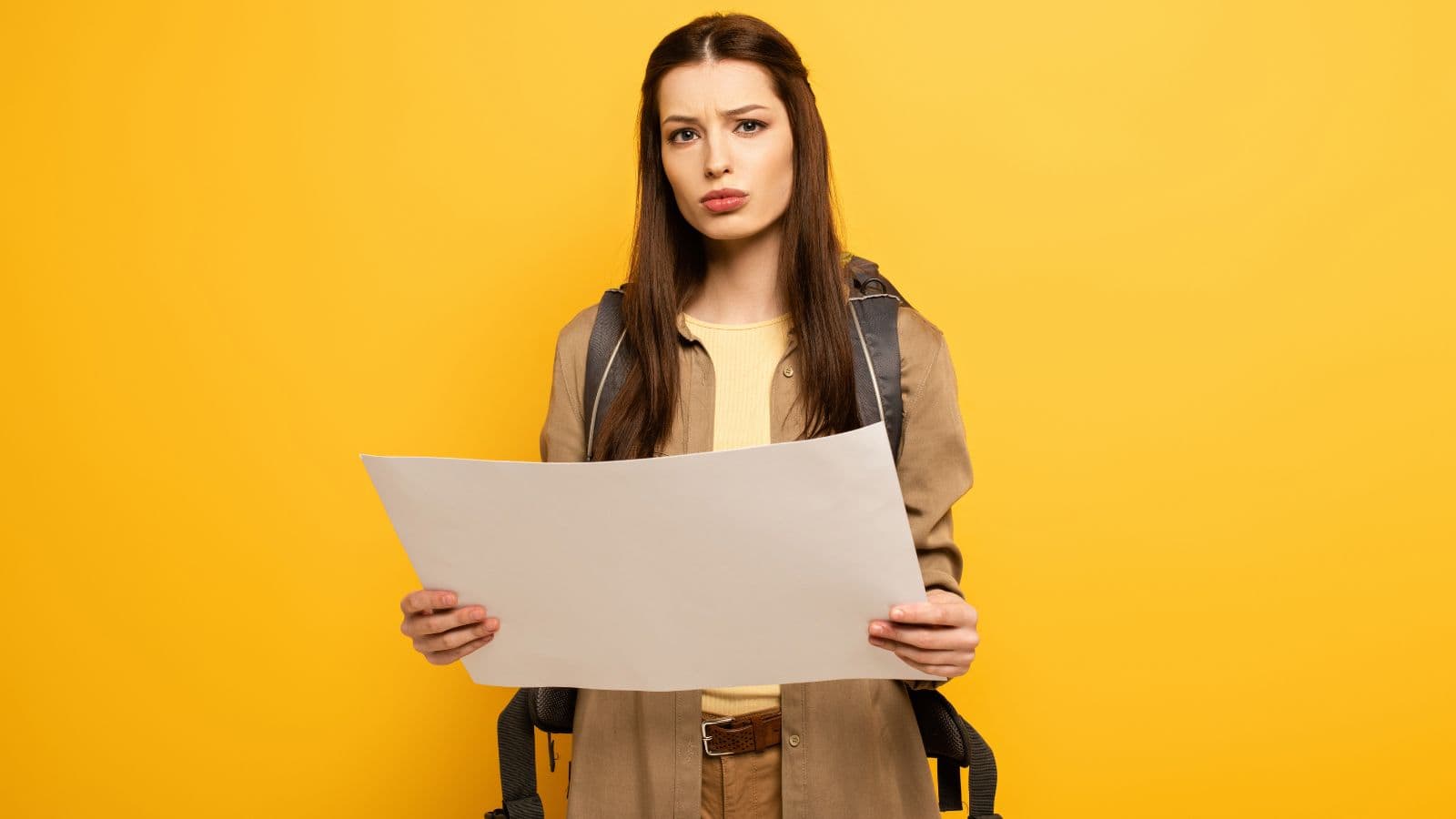 A young woman with long brown hair wearing a beige coat and backpack holds a large unfolded map, looking slightly confused, standing against a solid yellow background.