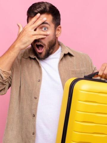 A man with a surprised expression holds a yellow suitcase in one hand while covering one eye with the other hand, standing in front of a solid pink background.