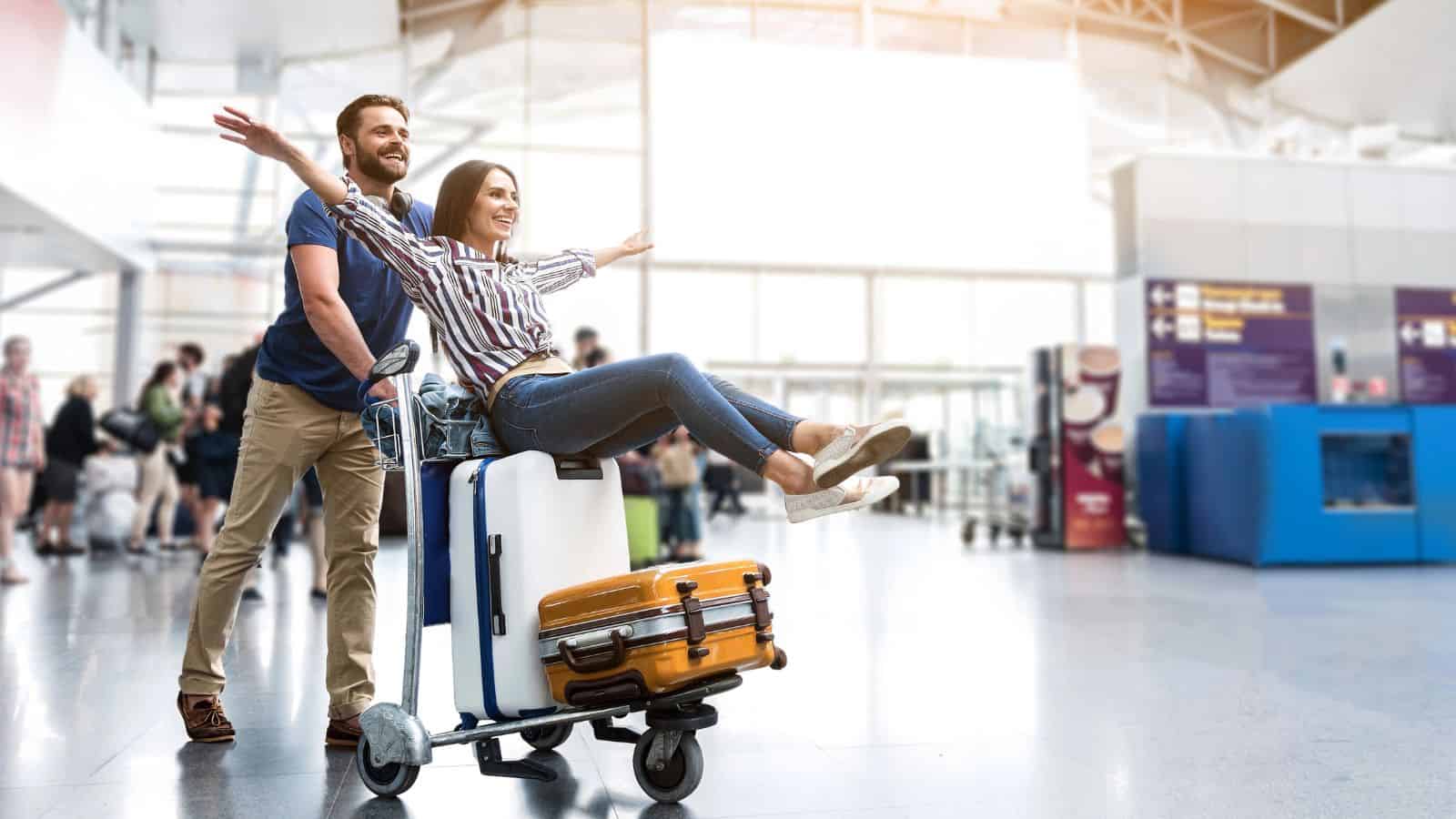 A man pushes a woman sitting on top of luggage on an airport trolley. The woman has her arms outstretched and is smiling, while the man smiles behind her. They are indoors with other travelers in the background.