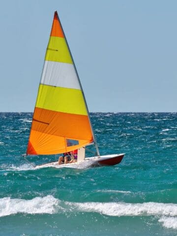 A small sailboat with a yellow, orange, and white sail moves across the ocean on a sunny day. Two people are on board, and another sailboat is visible in the distance. Waves are in the foreground.