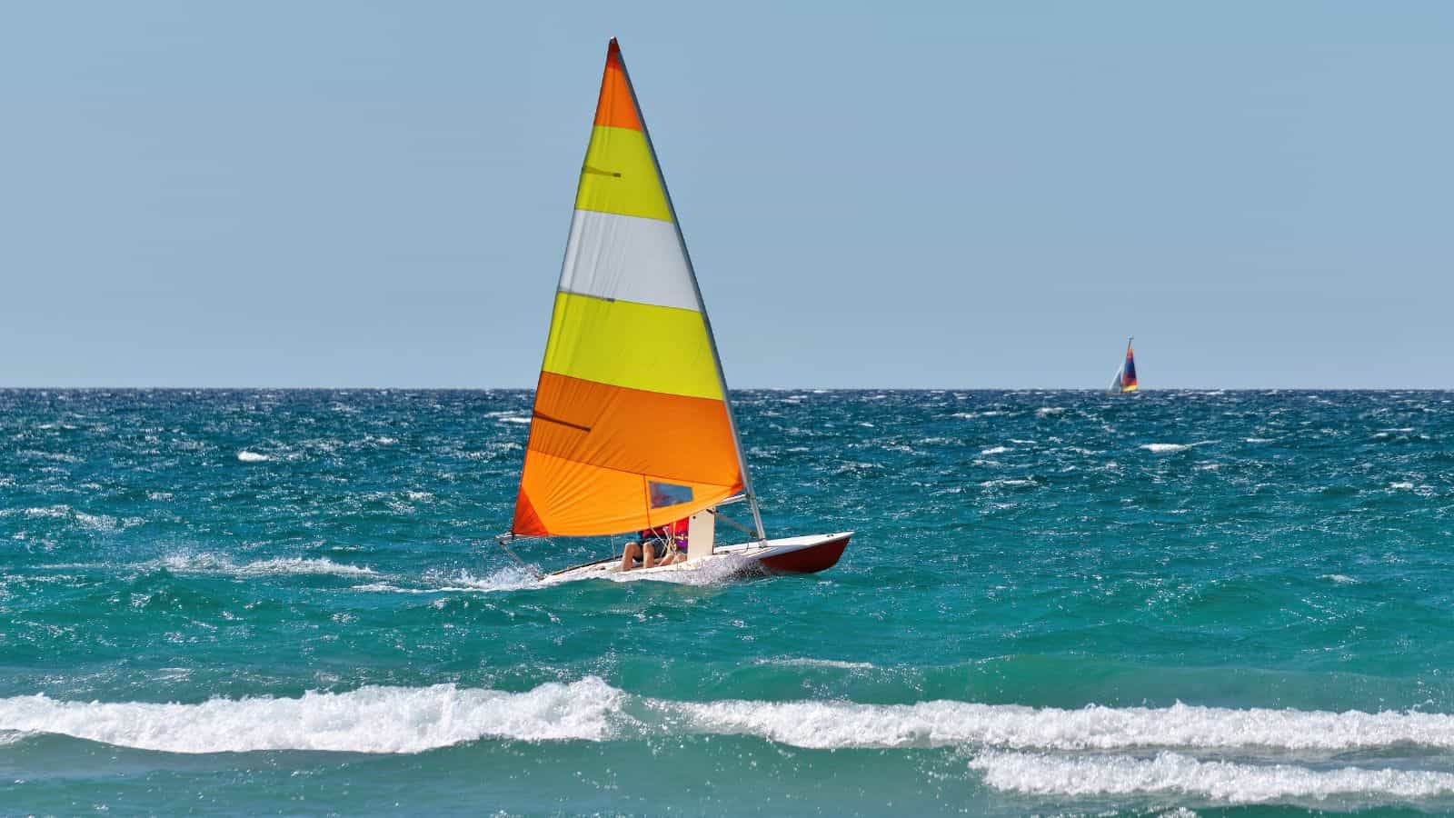 A small sailboat with a yellow, orange, and white sail moves across the ocean on a sunny day. Two people are on board, and another sailboat is visible in the distance. Waves are in the foreground.