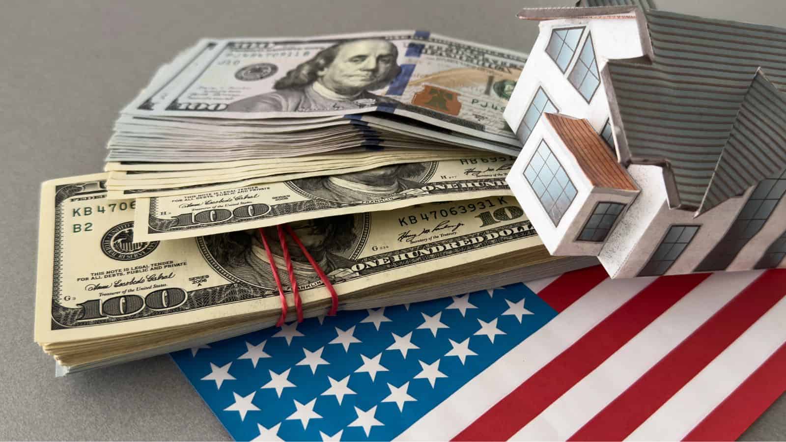A stack of US hundred-dollar bills, a small model house, and part of an American flag are placed on a gray surface.