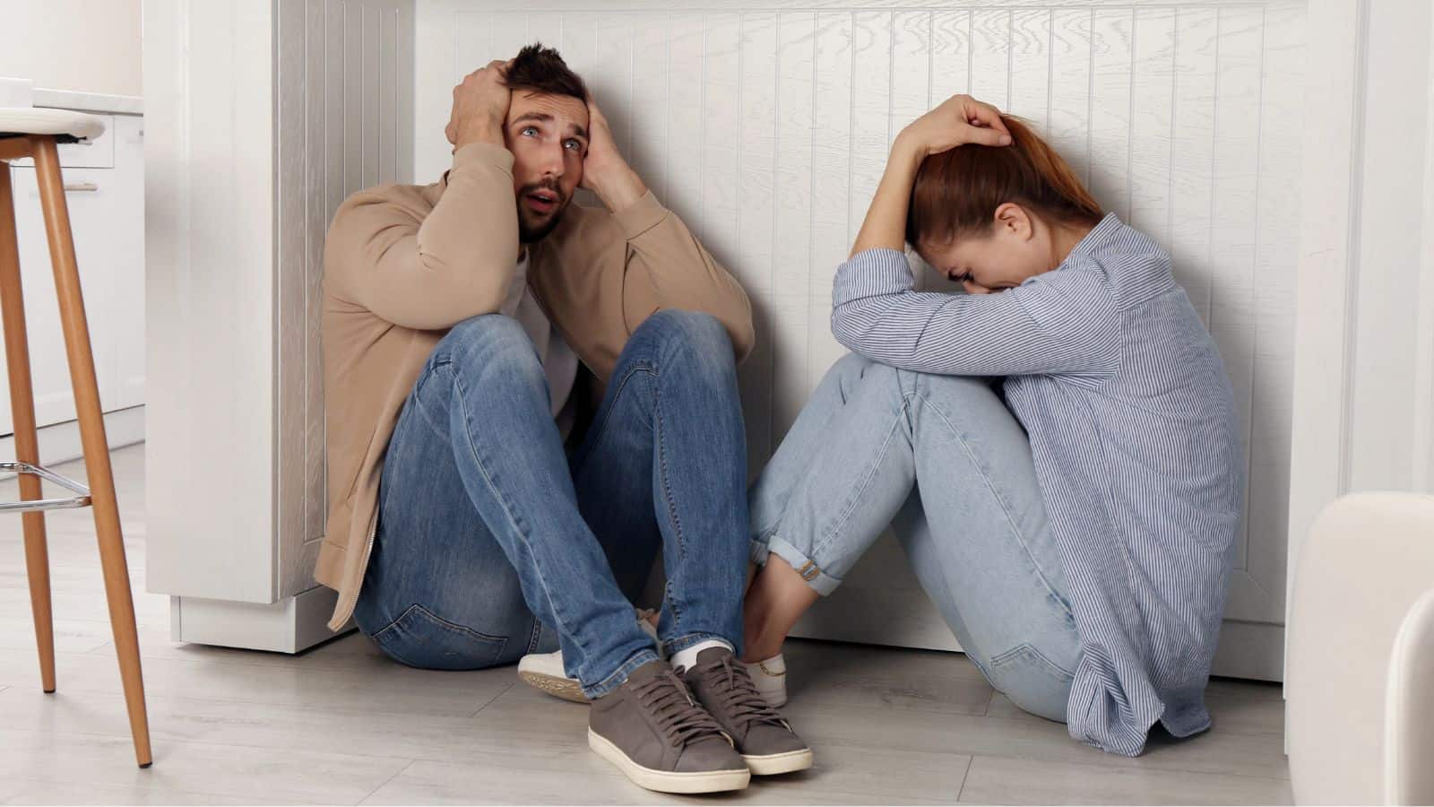 A man and a woman sit on the floor leaning against a white wall, both holding their heads with distressed expressions. They appear anxious or upset. The setting is a bright indoor space.