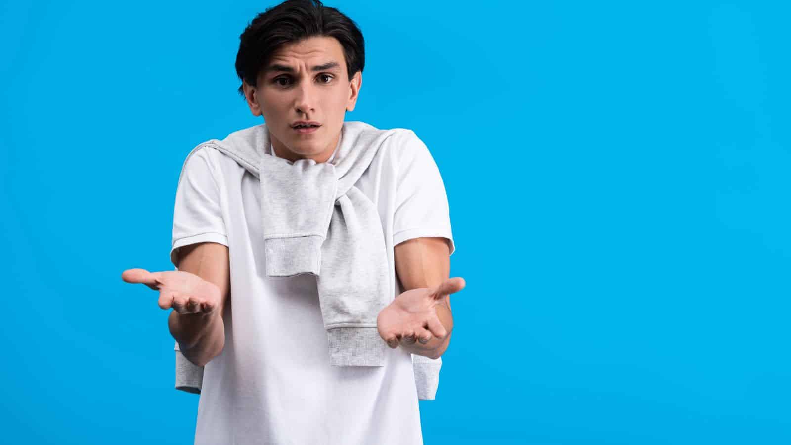 A young man in a white shirt with a gray sweatshirt draped over his shoulders stands against a blue background, holding his hands out in a confused or questioning gesture.