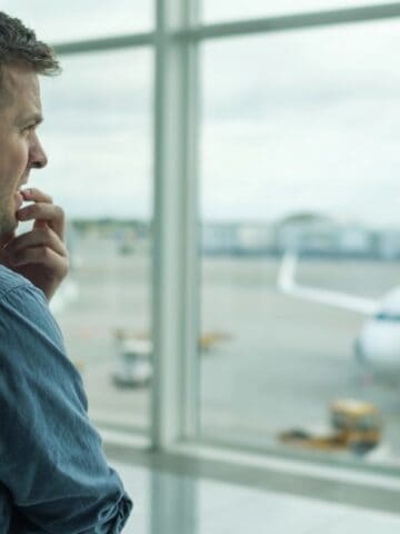 A man in a denim shirt stands by a large airport window, gazing thoughtfully at an airplane parked at the gate on a cloudy day, perhaps reflecting on travel tips for passengers facing a long flight experience.