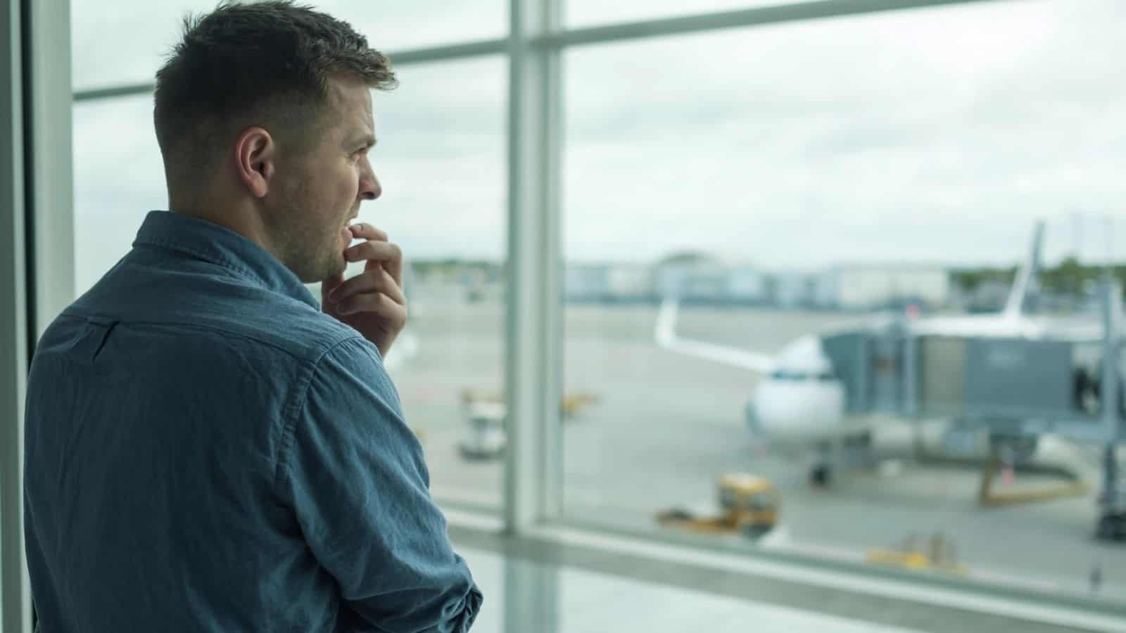 A man in a denim shirt stands by a large airport window, gazing thoughtfully at an airplane parked at the gate on a cloudy day, perhaps reflecting on travel tips for passengers facing a long flight experience.