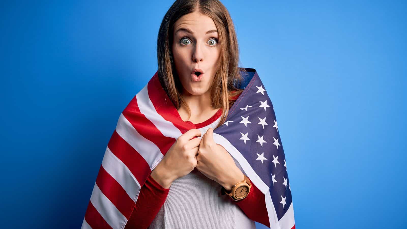 A woman with long brown hair stands in front of a blue background, wrapped in an American flag. She wears a white shirt and looks surprised, with her mouth forming an “O” shape.