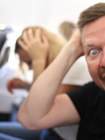 A man sits on an airplane seat with his mouth open wide and hands on his head, appearing shocked or scared—capturing the struggle many face with fear of flying. Behind him, a woman is bent over with her hands on her head as well.