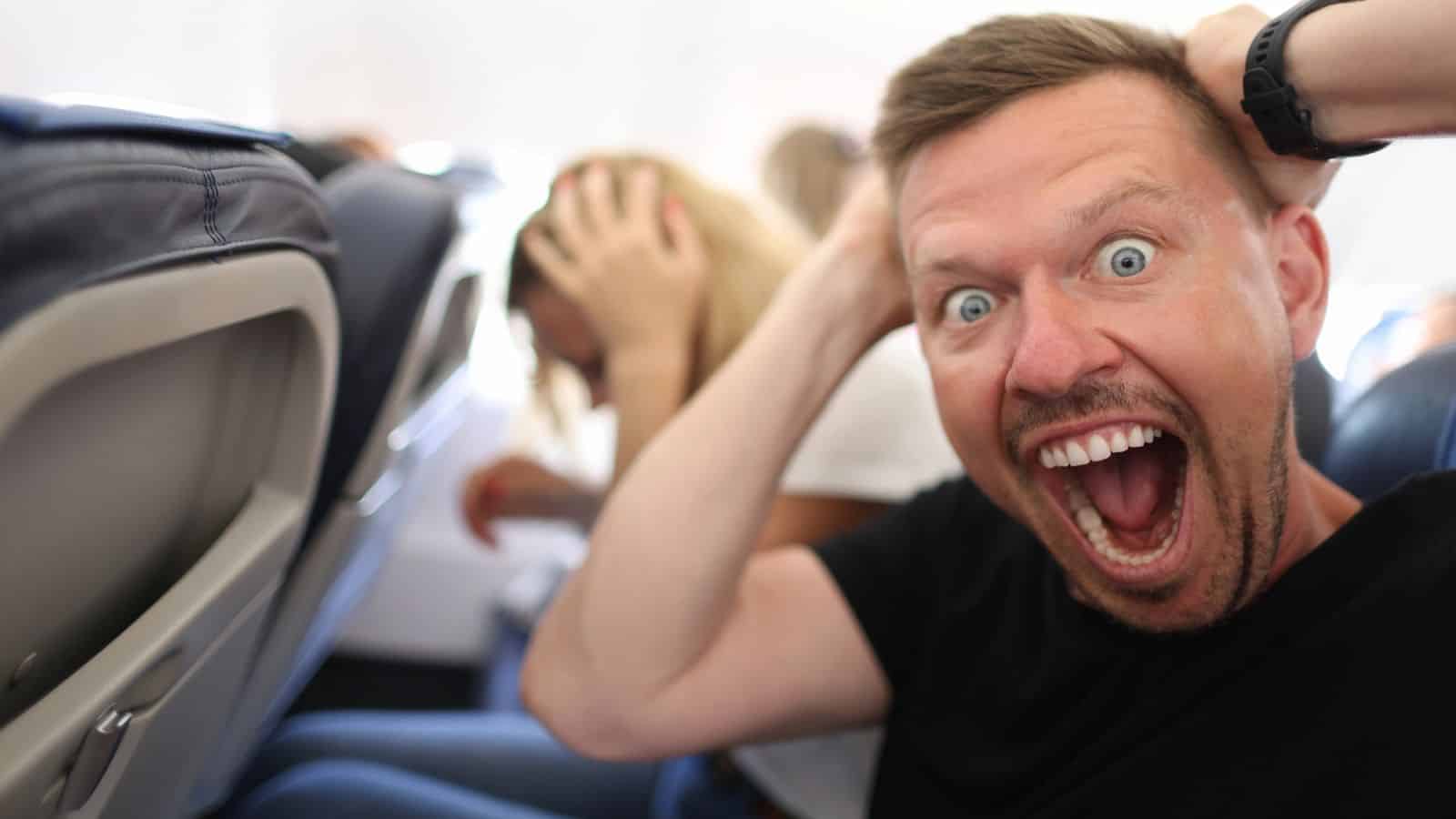 A man sits on an airplane seat with his mouth open wide and hands on his head, appearing shocked or scared&mdash;capturing the struggle many face with fear of flying. Behind him, a woman is bent over with her hands on her head as well.