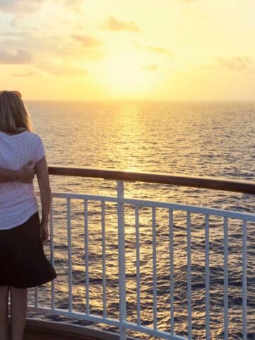 A couple stands on the deck of a ship, arm in arm, looking out at the ocean and sunset—a perfect scene for anyone seeking cruise tips or preparing for their first cruise. The sky has scattered clouds, and sunlight reflects on the water by the white railing.