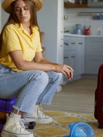 A woman wearing a yellow shirt, jeans, and a straw hat sits on an overpacked purple suitcase in a kitchen, looking frustrated—clearly facing some cruise packing mistakes. A closed red suitcase and a blue hat are on the floor nearby.