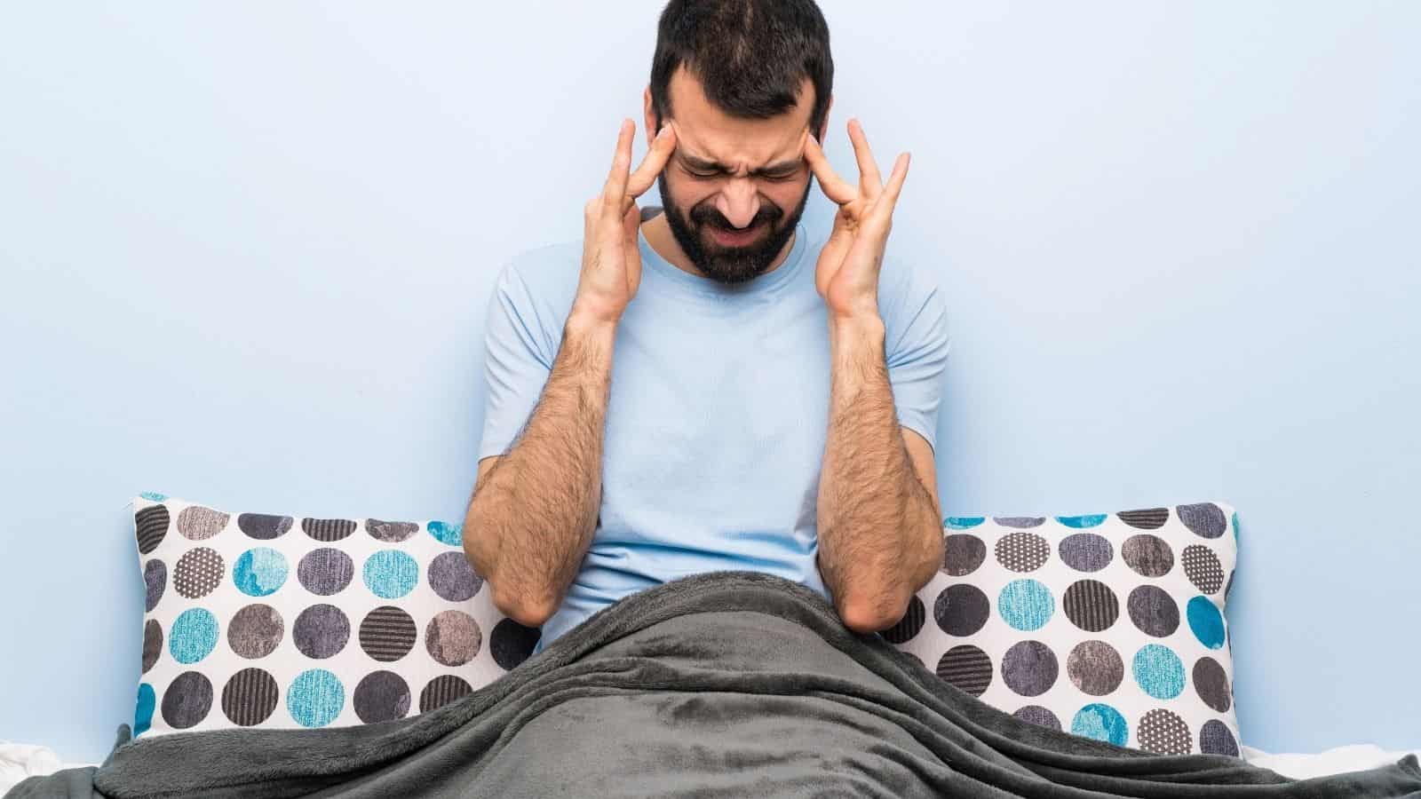 A man sits up in bed at one of the US hotel chains disappointing guests, with a blanket covering his lower body. He presses his fingers to his temples and closes his eyes in discomfort. Two patterned pillows are behind him.