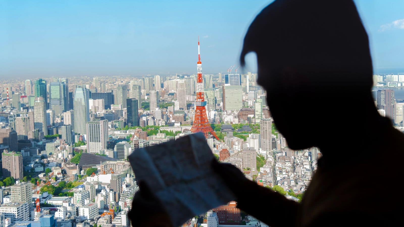 A person holding a map is silhouetted in the foreground, with a clear cityscape view of Tokyo and the Tokyo Tower prominently visible in the background during daytime.