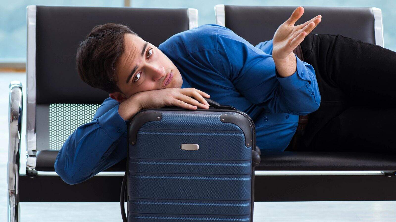 A man in a blue shirt lies on an airport bench, resting his head on a suitcase and raising one hand as if in frustration or confusion. He appears tired or bored while waiting.