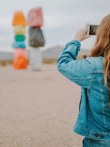 A person with long hair wearing a denim jacket takes a photo of brightly colored stacked rock sculptures in a desert landscape during daylight.