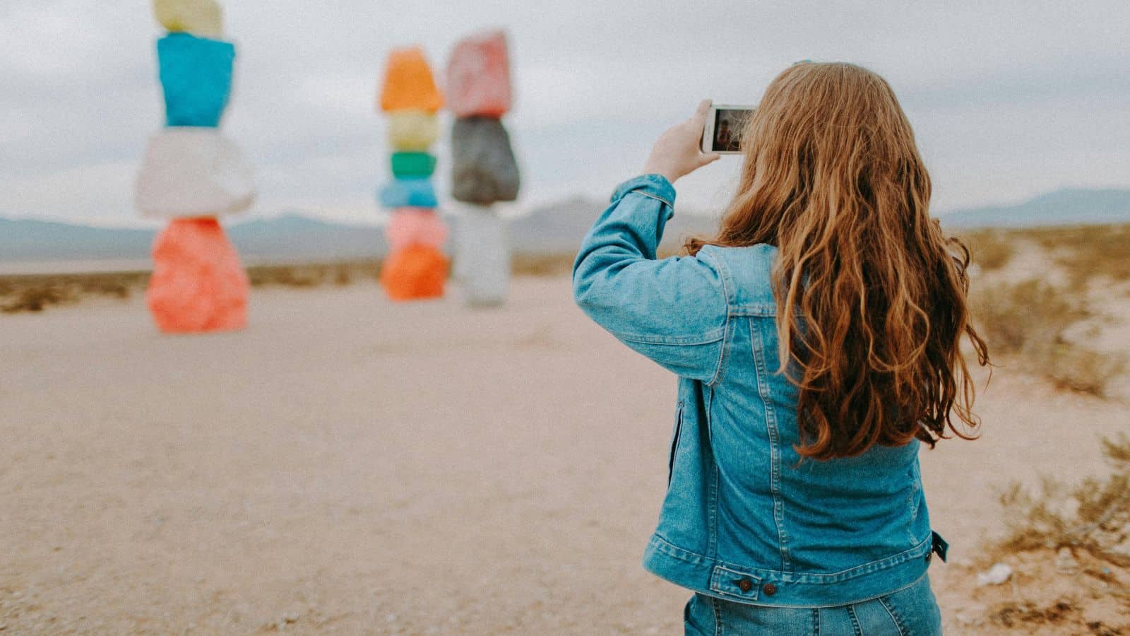 A person with long hair wearing a denim jacket takes a photo of brightly colored stacked rock sculptures in a desert landscape during daylight.