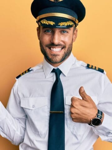 A man in a pilot uniform stands against a yellow background, smiling. He holds one hand out with the palm up and gives a thumbs-up with his other hand. He wears a hat, tie, and a wristwatch.