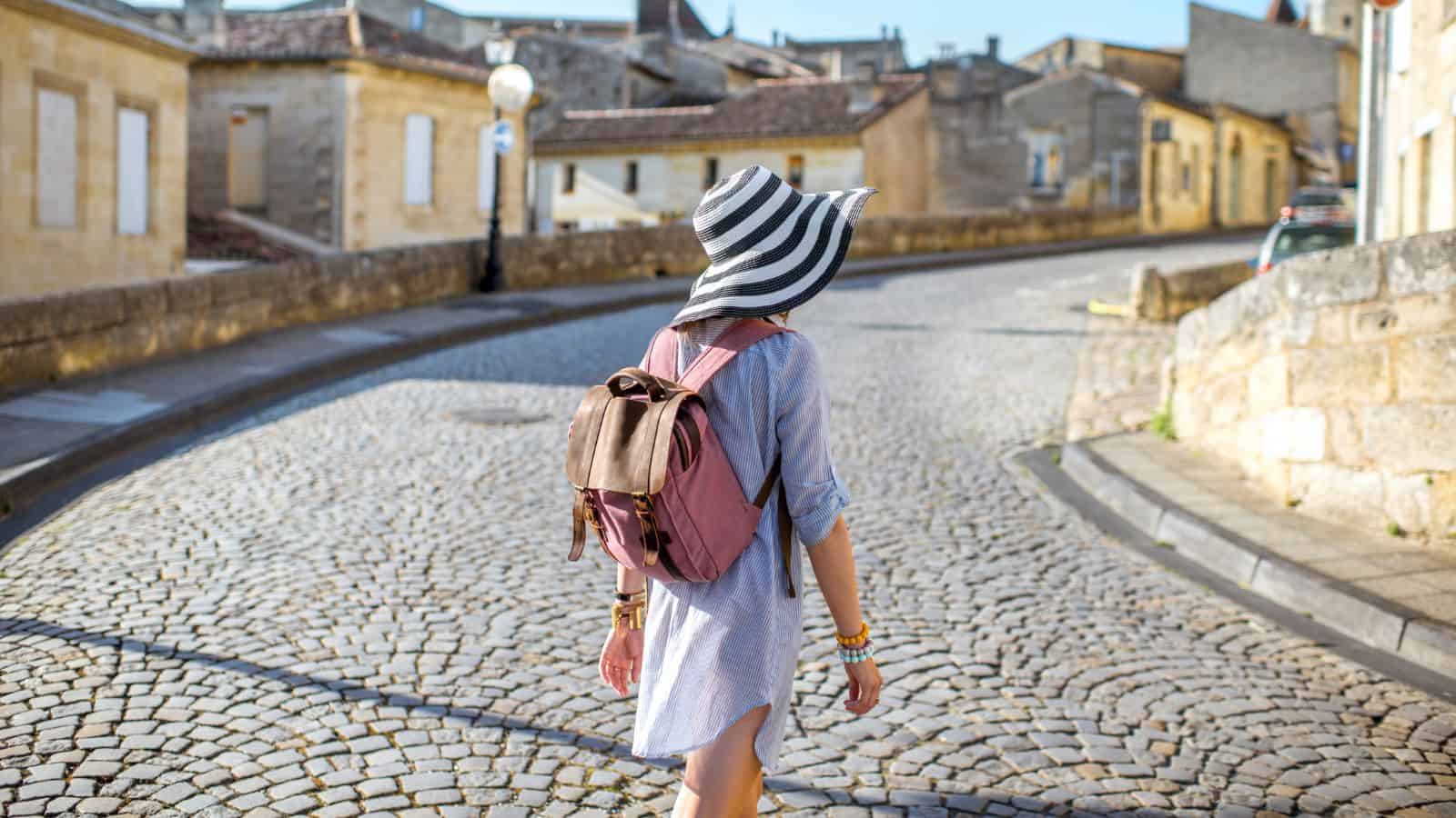 A person wearing a striped sunhat, blue dress, and pink backpack walks alone down a curved cobblestone street lined with old stone buildings on a sunny Europe travel day.