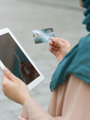 A person wearing a teal headscarf and beige clothing is holding a tablet in one hand and a credit card in the other, standing outdoors on a paved surface.