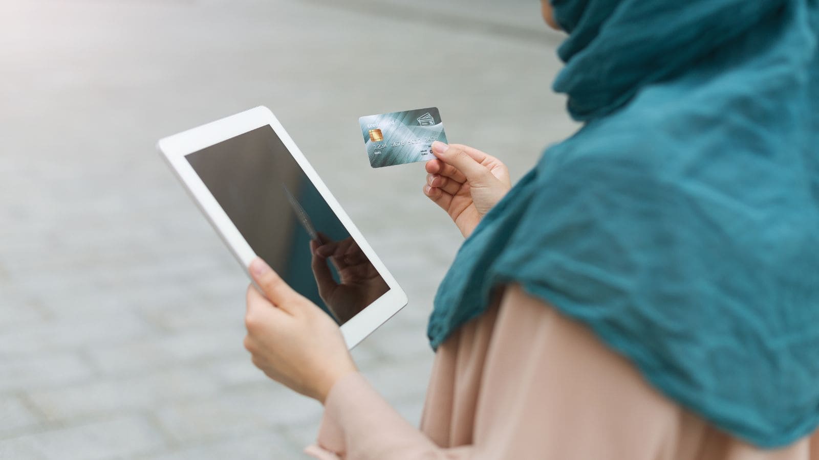 A person wearing a teal headscarf and beige clothing is holding a tablet in one hand and a credit card in the other, standing outdoors on a paved surface.