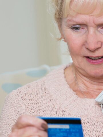 An older woman with blonde hair is sitting on a sofa, holding a landline phone to her ear with one hand and looking at a credit card in her other hand. She appears to be in conversation.