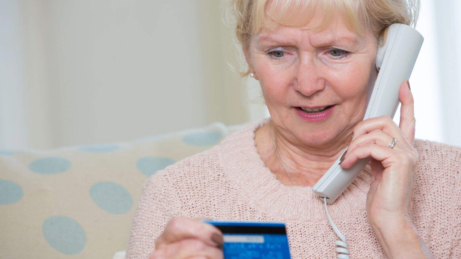 An older woman with blonde hair is sitting on a sofa, holding a landline phone to her ear with one hand and looking at a credit card in her other hand. She appears to be in conversation.