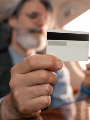 A man with gray hair and beard holds a credit card in one hand and a smartphone in the other, sitting at a table in what appears to be the cabin of a private jet.