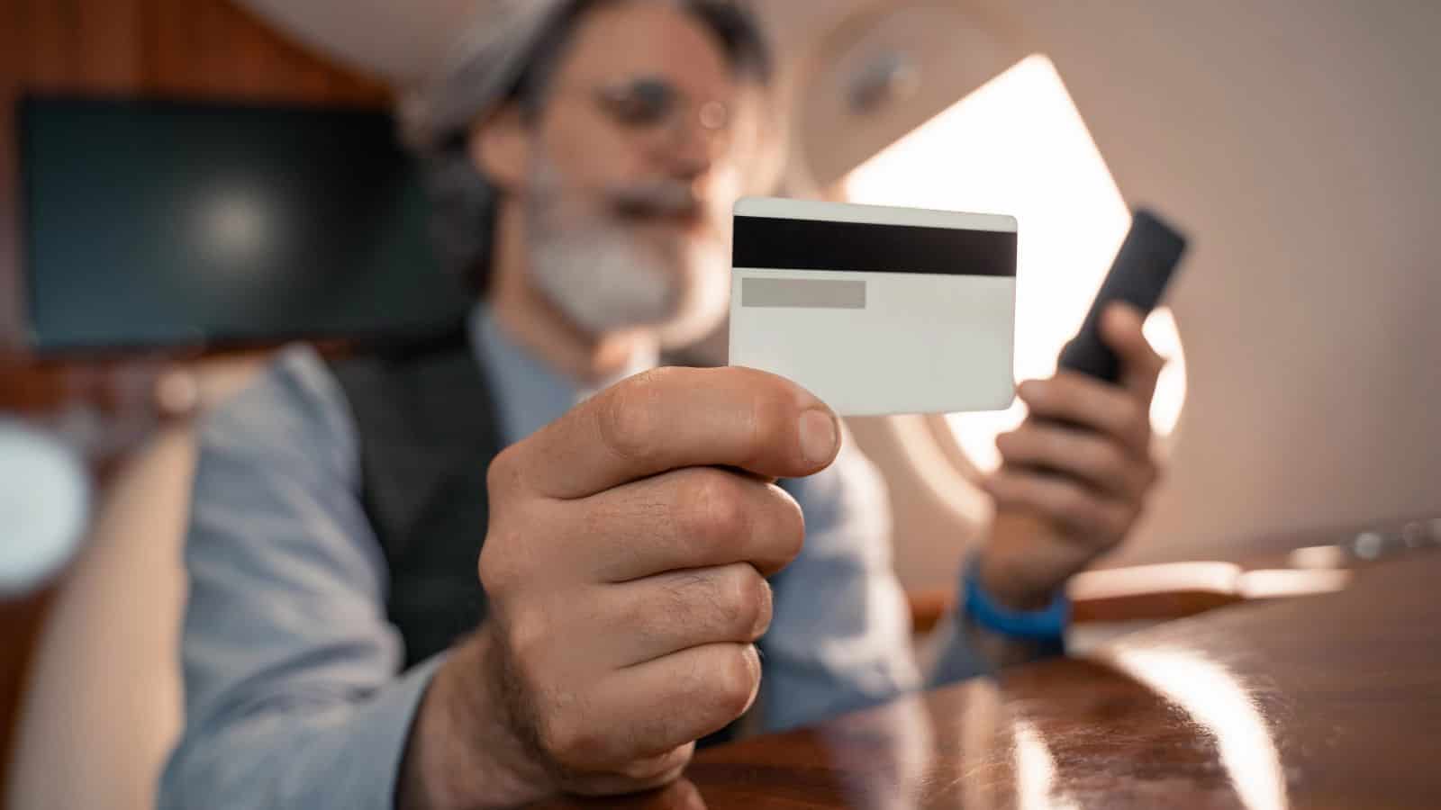A man with gray hair and beard holds a credit card in one hand and a smartphone in the other, sitting at a table in what appears to be the cabin of a private jet.