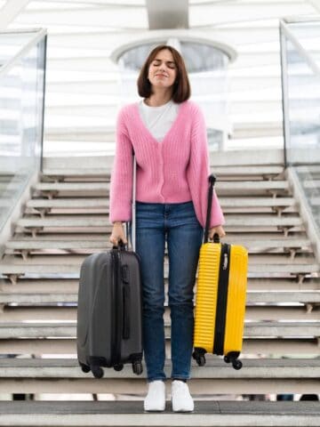 A woman in a pink sweater and jeans stands on stairs holding a gray suitcase in one hand and a yellow suitcase in the other, with glass railings and a covered structure in the background.