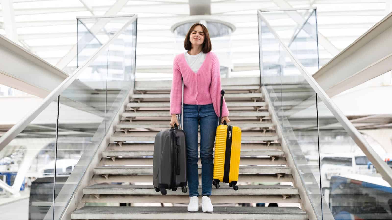 A woman in a pink sweater and jeans stands on stairs holding a gray suitcase in one hand and a yellow suitcase in the other, with glass railings and a covered structure in the background.