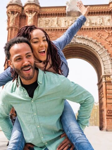 A man gives a woman a piggyback ride in front of a large brick archway. Both are smiling, and the woman has her arms outstretched. The scene takes place outdoors in a city square with people and trees in the background.
