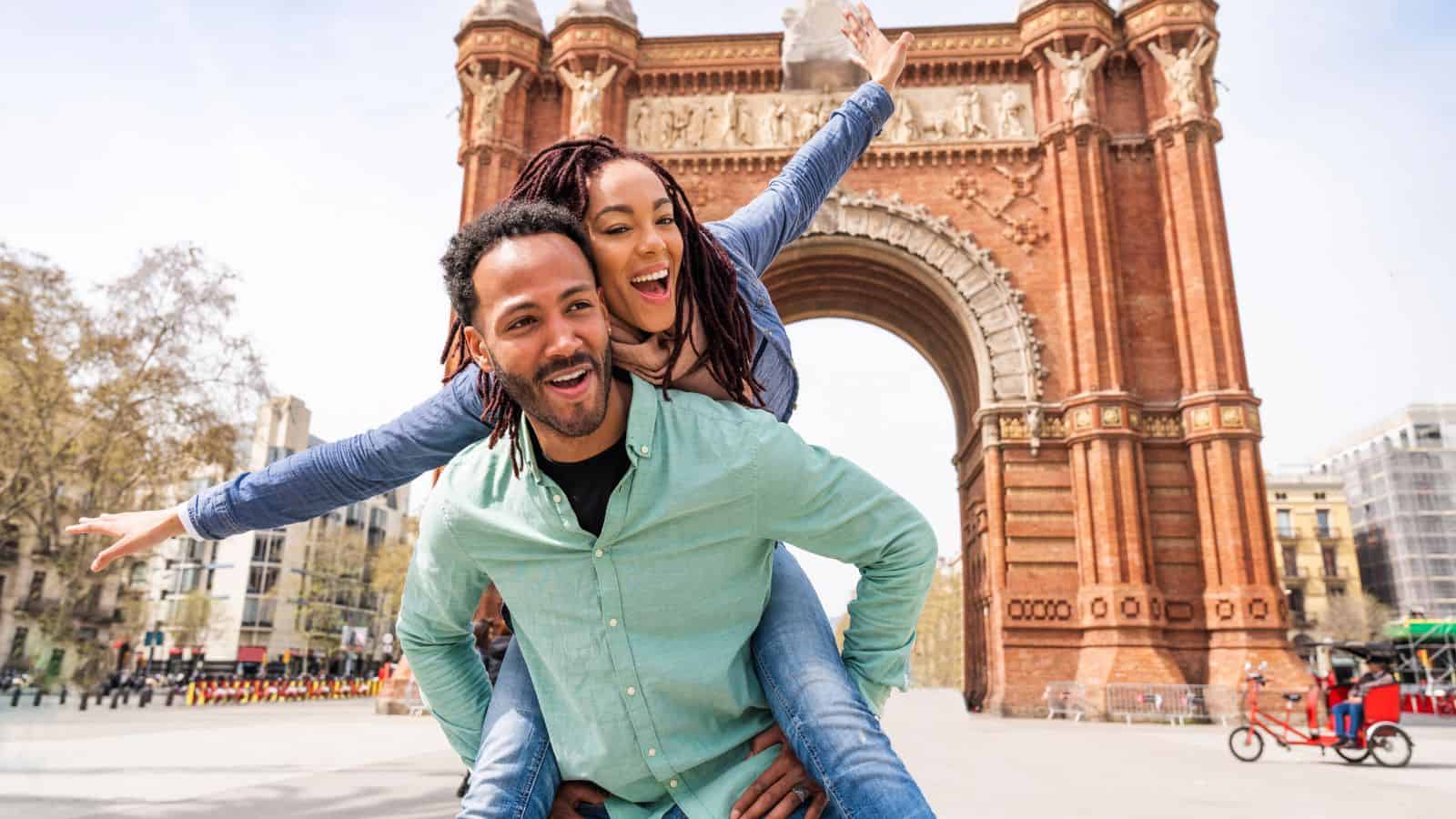 A man gives a woman a piggyback ride in front of a large brick archway. Both are smiling, and the woman has her arms outstretched. The scene takes place outdoors in a city square with people and trees in the background.