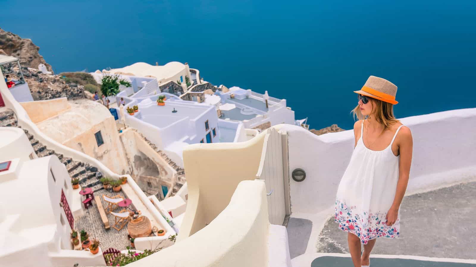 A woman in a white sundress and orange hat stands on a sunlit path with white buildings and terraces overlooking the blue sea in a coastal Mediterranean village.