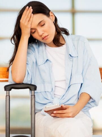 A woman sits on a bench indoors with her eyes closed, resting her head on one hand. She holds a smartphone in her other hand and has a suitcase beside her. The setting appears to be an airport or station.