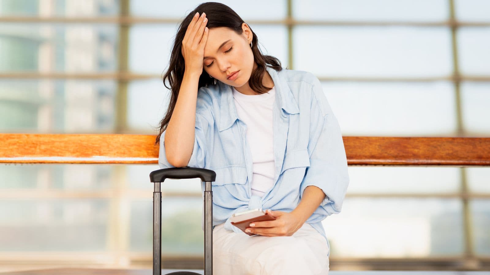 A woman sits on a bench indoors with her eyes closed, resting her head on one hand. She holds a smartphone in her other hand and has a suitcase beside her. The setting appears to be an airport or station.
