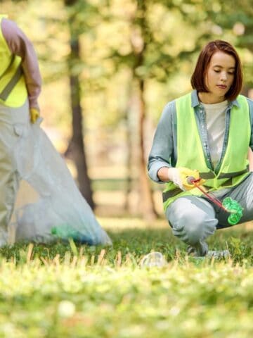 Two people wearing yellow safety vests pick up litter in a grassy park area, using grabbers and placing trash into large blue garbage bags. Trees and greenery are visible in the background.