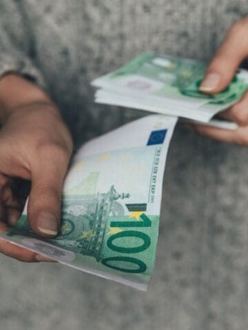 A person holding and counting several 100 euro banknotes with both hands, wearing a gray long-sleeved garment.