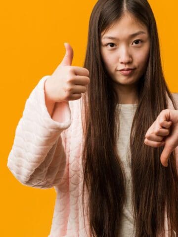 A young woman with long hair wearing a light pink textured jacket stands against an orange background, giving a thumbs up with one hand and a thumbs down with the other.