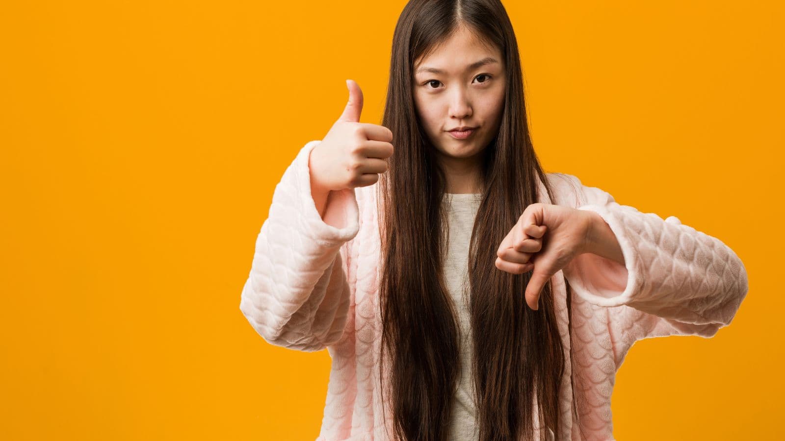 A young woman with long brown hair stands in front of an orange background, wearing a light pink jacket. She is giving a thumbs up with her right hand and a thumbs down with her left hand.
