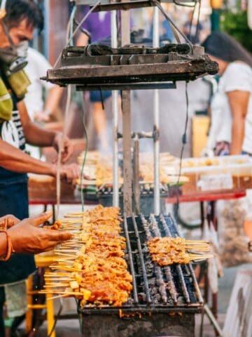 Several people are grilling and selling skewers of meat at a busy outdoor street food stall. Smoke rises from the grill as customers and vendors, some wearing masks, interact in the crowded scene.