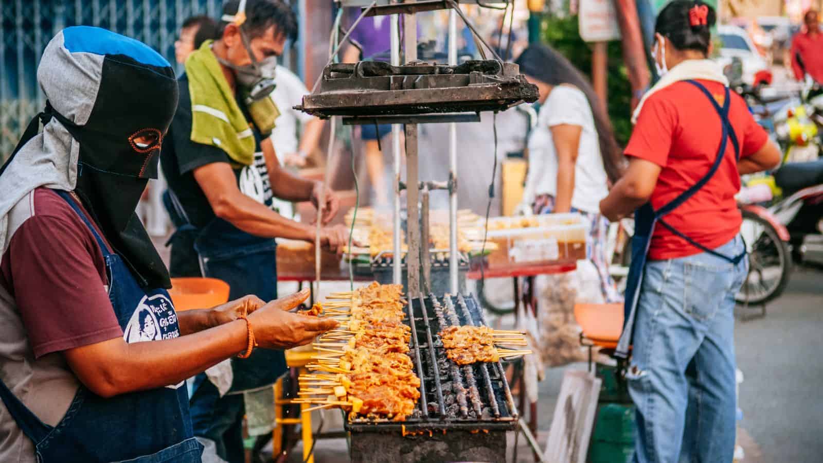Several people are grilling and selling skewers of meat at a busy outdoor street food stall. Smoke rises from the grill as customers and vendors, some wearing masks, interact in the crowded scene.