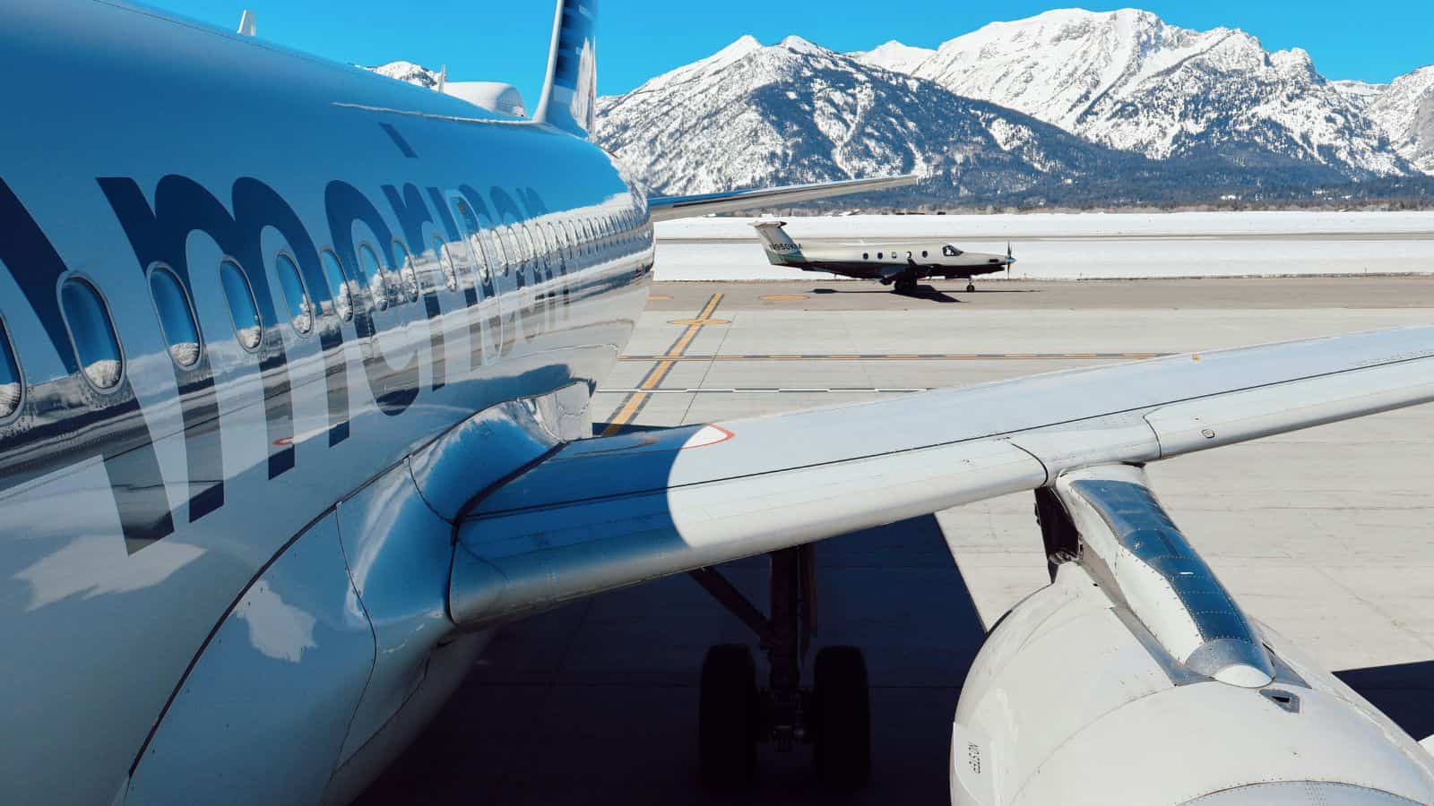 A close-up view of an American Airlines plane on an airport tarmac, with a small jet in the background and snow-covered mountains under a clear blue sky.