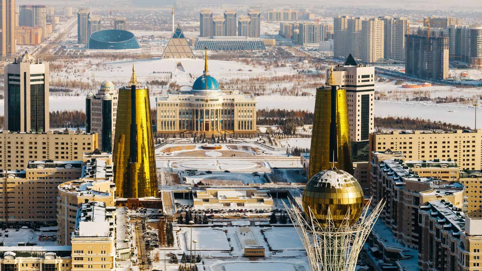 A cityscape of Astana, Kazakhstan, showing modern buildings with golden towers, the blue-domed Ak Orda Presidential Palace, snowy streets, and the distinctive egg-shaped Bayterek Tower in the foreground.
