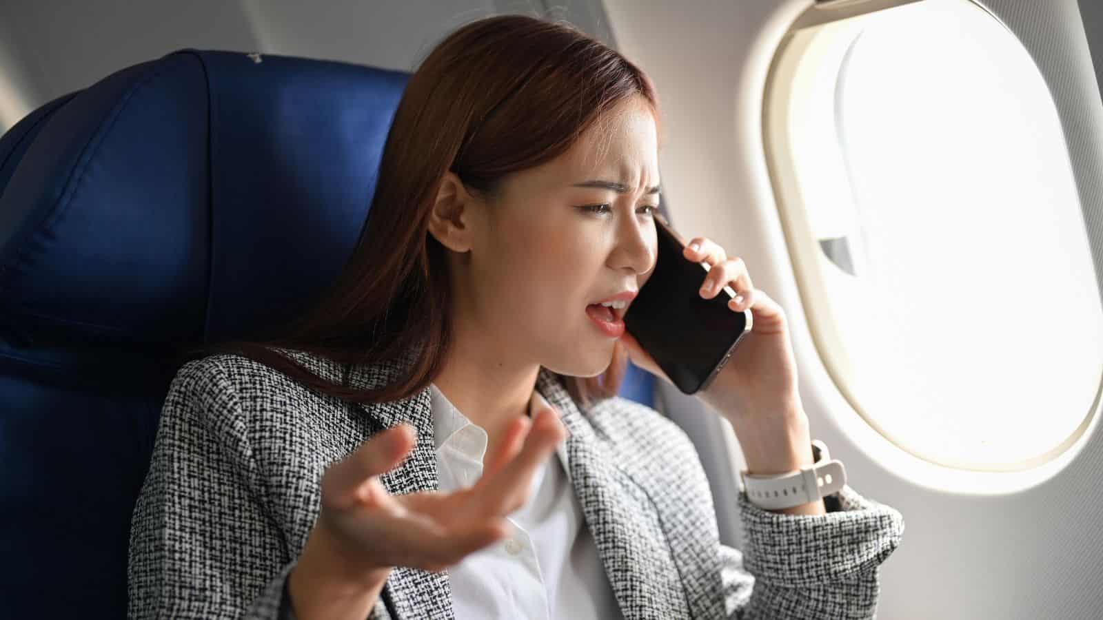 A woman in business attire sits on an airplane, holding a phone to her ear and gesturing with her other hand, appearing frustrated or upset. She is seated by the window.