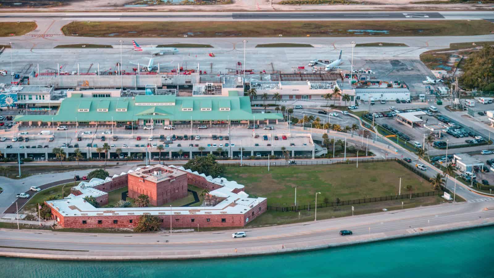 Aerial view of an airport terminal with several airplanes on the tarmac, a historic red brick fort in the foreground, a road along the waterfront, and parking lots with cars.