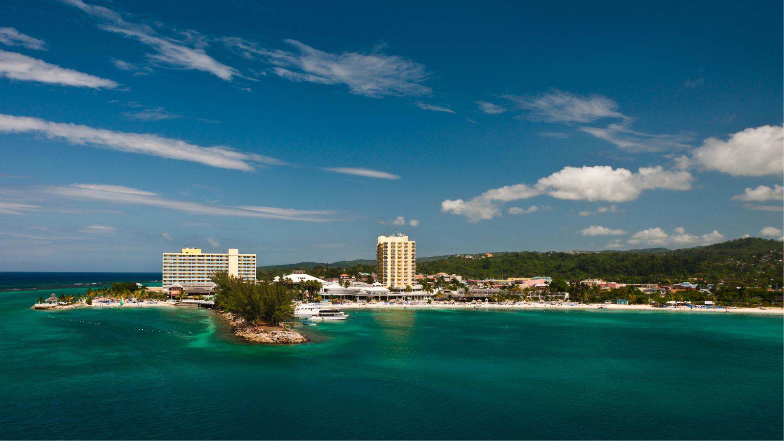 A coastal scene with turquoise water, two tall buildings near the shore, a small peninsula with trees, and hills in the background under a blue sky with scattered clouds.